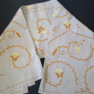 May include: A pair of off-white linen table runners with embroidered floral designs in shades of yellow and brown. The floral pattern features stylized leaves and flowers.