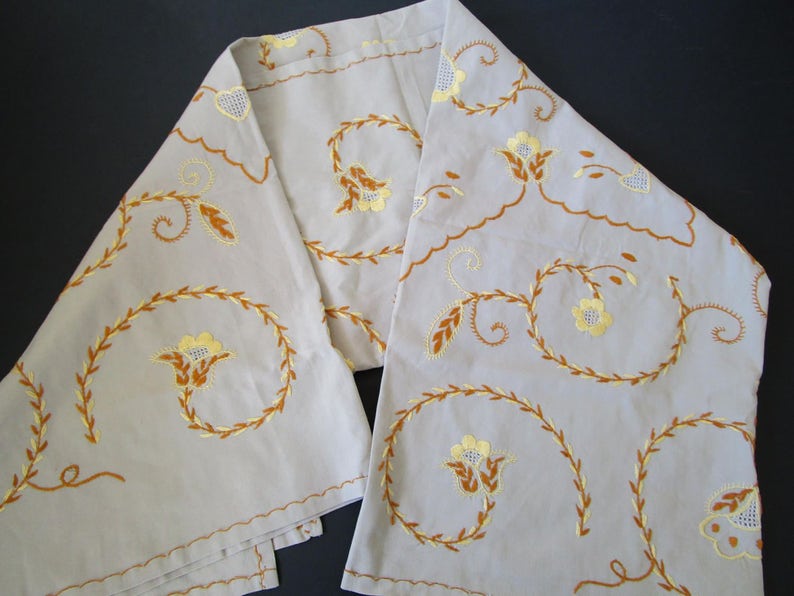 May include: A pair of off-white linen table runners with embroidered floral designs in shades of yellow and brown. The floral pattern features stylized leaves and flowers.
