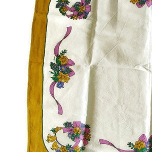 May include: A white fabric square with a gold border and floral designs. The floral designs are wreaths of yellow, pink, and purple flowers with green leaves.