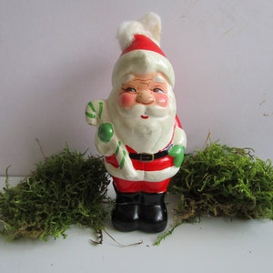May include: A vintage Santa Claus figurine with a red and white hat, red suit, and black boots. The figurine holds a green and white striped candy cane. The Santa has a white beard and rosy cheeks. A black belt with a silver buckle is around his waist.