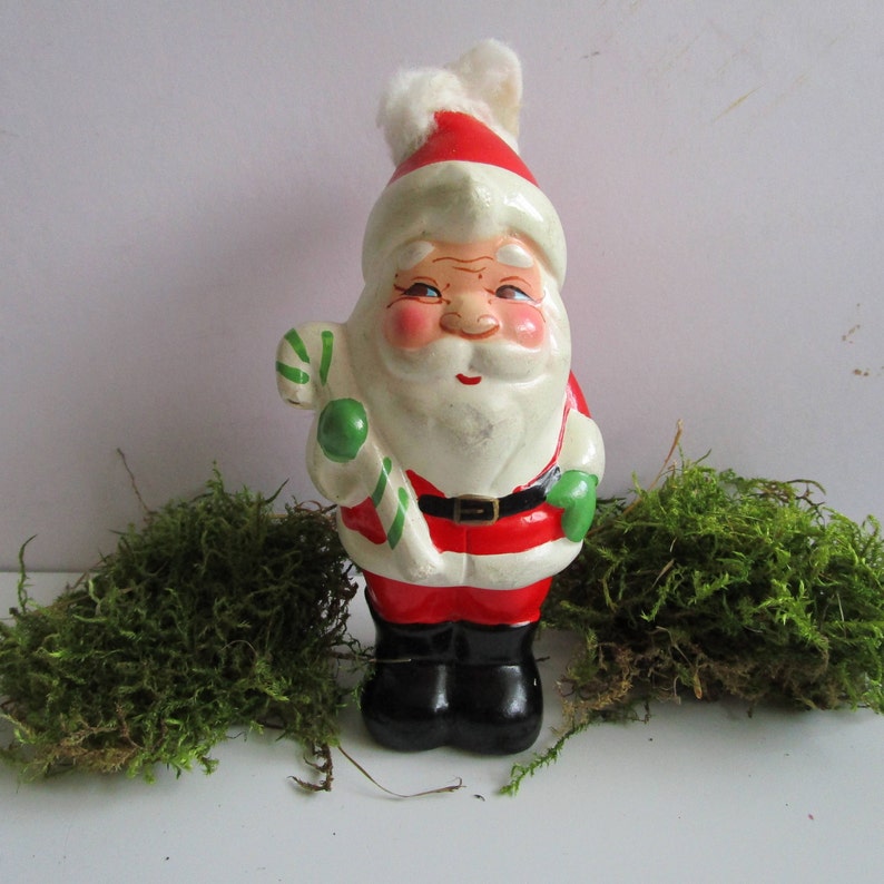 May include: A vintage Santa Claus figurine with a red and white hat, red suit, and black boots. The figurine holds a green and white striped candy cane. The Santa has a white beard and rosy cheeks. A black belt with a silver buckle is around his waist.