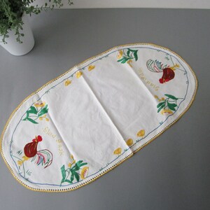 May include: White oval table runner with a yellow crocheted border. The runner features embroidered roosters, flowers, and the text "Glad P&aring;sk" and "Sista &Auml;gget".