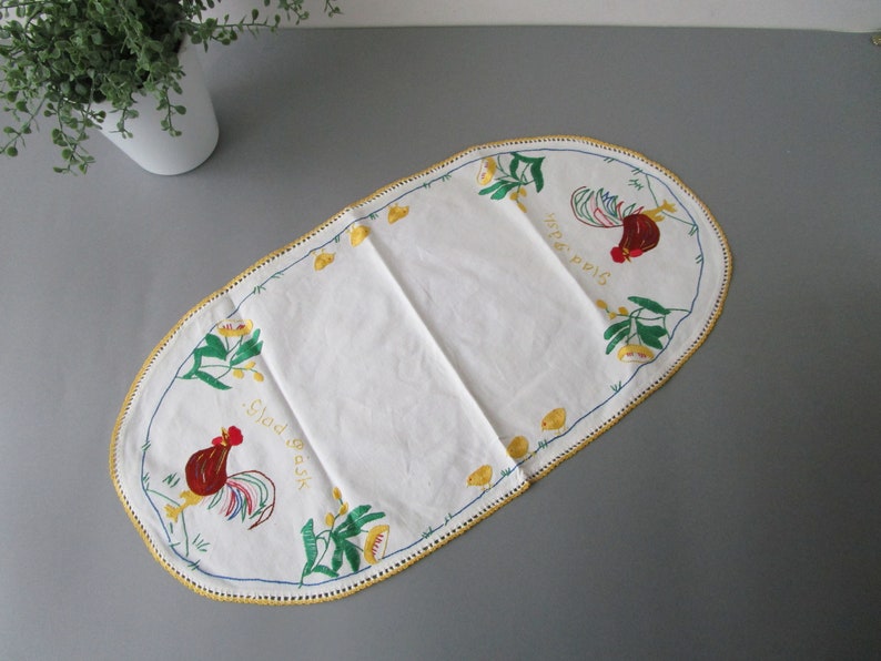 May include: White oval table runner with a yellow crocheted border. The runner features embroidered roosters, flowers, and the text "Glad P&aring;sk" and "Sista &Auml;gget".