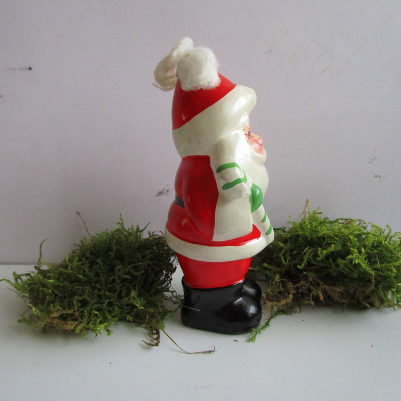 May include: A vintage Santa Claus figurine, painted in red, white, and black. The figurine has a white beard, a red suit, and black boots. It is holding a green and white striped candy cane. The figurine is a Christmas decoration.