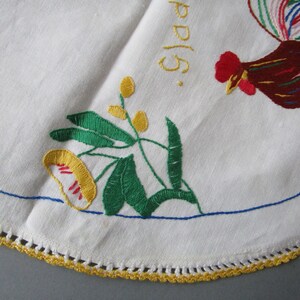 May include: White embroidered tablecloth with a rooster, flowers, and leaves. The tablecloth has a yellow crocheted border.