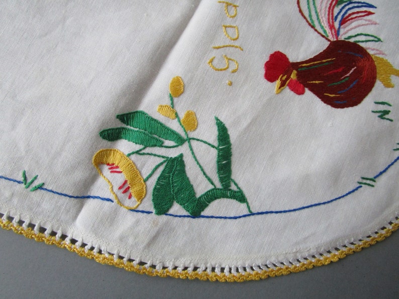 May include: White embroidered tablecloth with a rooster, flowers, and leaves. The tablecloth has a yellow crocheted border.