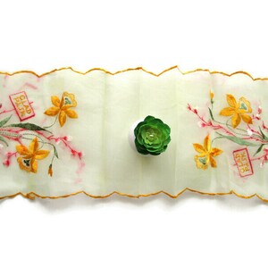 May include: A rectangular, off-white sheer table runner with scalloped edges trimmed in yellow thread. It features embroidered yellow daffodil flowers, green foliage, and pink accents. Two red rectangular boxes contain the text GLAD PASK.