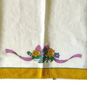 May include: A white fabric tablecloth with a yellow border and a pink ribbon with a floral design. The flowers are yellow, purple, and green.