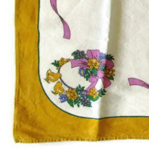 May include: A white fabric with a yellow border and a floral design featuring a pink bow, yellow flowers, and birds. The fabric has a scalloped edge.