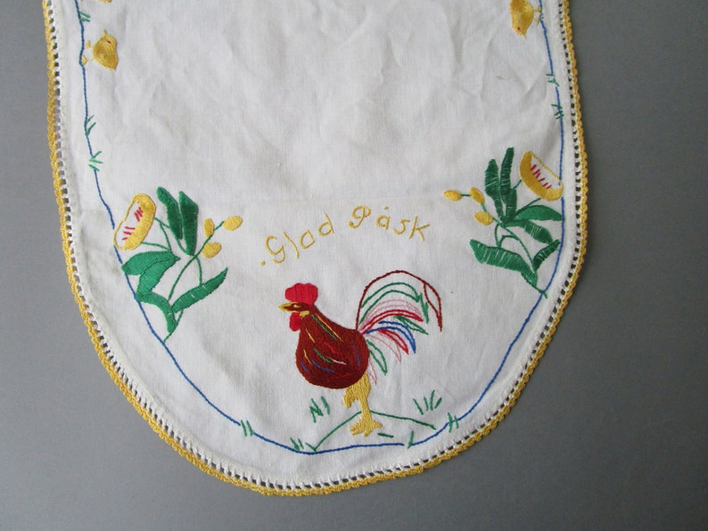 May include: White oval tablecloth with a yellow crocheted border. The tablecloth is embroidered with a rooster, flowers, and the text "Glad P&aring;sk" in yellow thread.