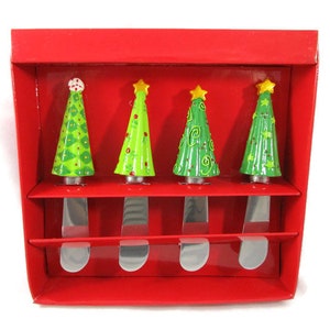 Peut inclure: Un ensemble de quatre couteaux à tartiner en métal avec des manches en forme de sapins de Noël verts, chacun arborant des motifs uniques et une étoile jaune au sommet. Ils sont présentés dans une boîte en carton rouge avec des fentes.