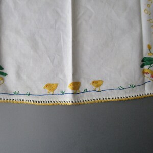 May include: White linen tablecloth with yellow and green embroidered floral designs and a rooster with red and yellow accents. The text "Glad Tidings" is embroidered in the top left corner. Three yellow chicks are embroidered at the bottom of the tablecloth.
