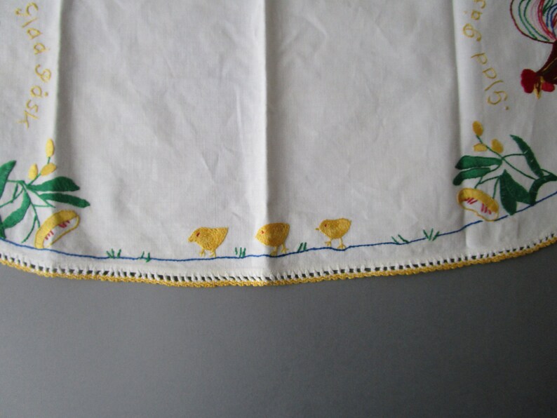May include: White linen tablecloth with yellow and green embroidered floral designs and a rooster with red and yellow accents. The text "Glad Tidings" is embroidered in the top left corner. Three yellow chicks are embroidered at the bottom of the tablecloth.