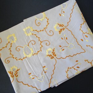 May include: A white tablecloth with embroidered floral designs in yellow and brown. The tablecloth has a scalloped edge.