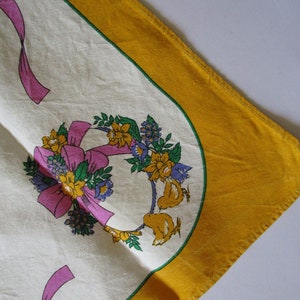 May include: A white tablecloth with a yellow border and a floral design featuring pink ribbons, yellow flowers, and green leaves. Two small yellow birds are perched on the floral wreath.