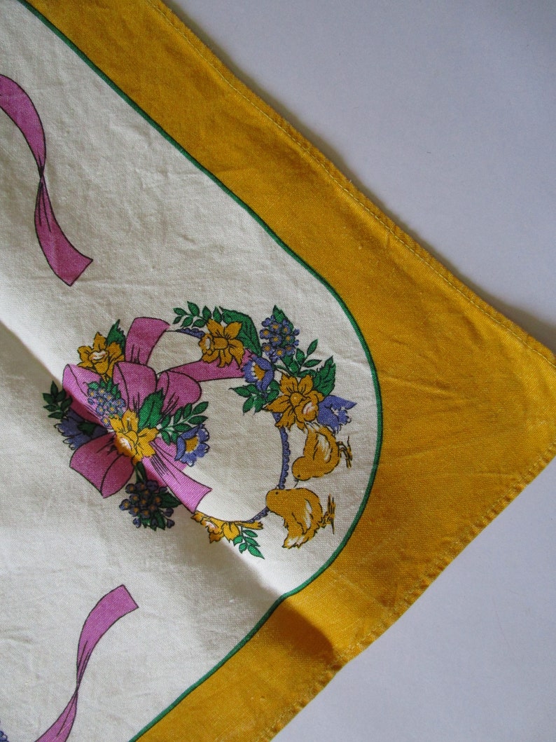 May include: A white tablecloth with a yellow border and a floral design featuring pink ribbons, yellow flowers, and green leaves. Two small yellow birds are perched on the floral wreath.
