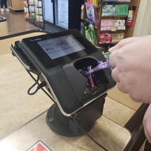 May include: A black and silver credit card reader with a screen displaying the text "Please Wait For Cashier" and the logo for Sprouts Farmers Market. The screen also shows the Apple Pay logo. A purple and silver tool is being used to touch the reader.