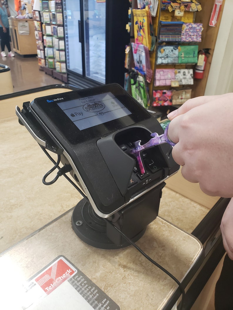 May include: A black and silver credit card reader with a screen displaying the text "Please Wait For Cashier" and the logo for Sprouts Farmers Market. The screen also shows the Apple Pay logo. A purple and silver tool is being used to touch the reader.