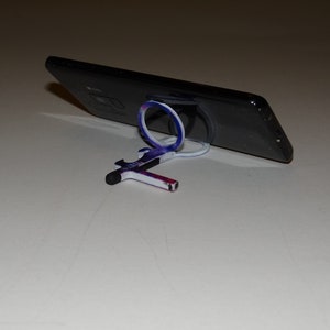 May include: A black smartphone with a white and blue phone ring stand. The phone is propped up on the stand, and the stand is on a white surface.