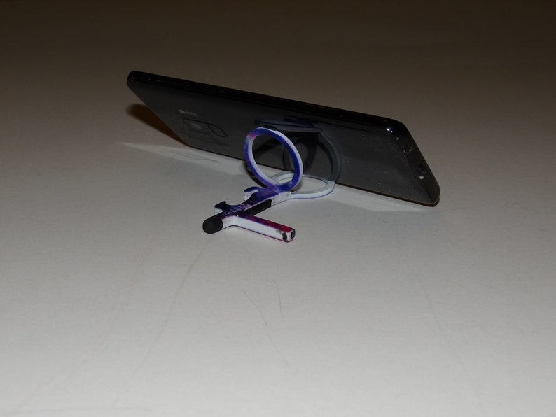 May include: A black smartphone with a white and blue phone ring stand. The phone is propped up on the stand, and the stand is on a white surface.