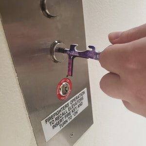 May include: A silver metal panel with a red button labelled "Bypass" and "Fire Service". The button has a keyhole and is labelled "Off" and "On". A sticker on the panel reads "Firefighters' Operation to Recall Elevators Insert Fire Key and Turn to "On"". A hand is holding a purple and black tool with the word "Awake" printed on it, and is inserting the tool into the keyhole.