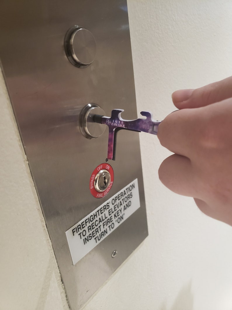 May include: A silver metal panel with a red button labelled "Bypass" and "Fire Service". The button has a keyhole and is labelled "Off" and "On". A sticker on the panel reads "Firefighters' Operation to Recall Elevators Insert Fire Key and Turn to "On"". A hand is holding a purple and black tool with the word "Awake" printed on it, and is inserting the tool into the keyhole.