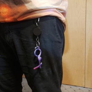 May include: A black retractable keychain with a purple and blue key-shaped tool attached. The tool has a loop at the top and a pointed end.