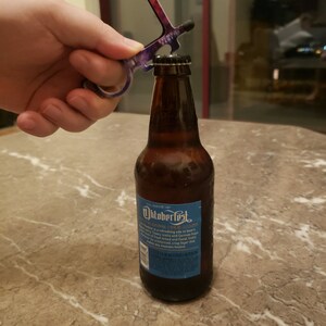May include: A purple and black multi-tool bottle opener is being used to open a brown glass bottle of Oktoberfest beer. The bottle label is blue and features the text "Oktoberfest" and "German Style Lager".