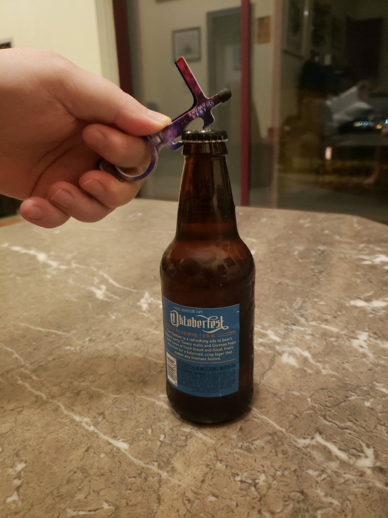 May include: A purple and black multi-tool bottle opener is being used to open a brown glass bottle of Oktoberfest beer. The bottle label is blue and features the text "Oktoberfest" and "German Style Lager".