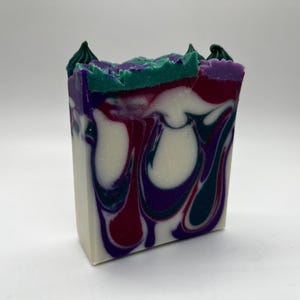 May include: A rectangular bar of handmade soap with a layered design. The soap features swirls of white, deep red, teal, and purple. The top edge has a textured, jagged appearance with green and purple accents.