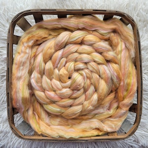 May include: A braided bundle of colorful silk fibers in shades of peach, pink, yellow, and green. The fibers are arranged in a spiral pattern and are contained within a wooden basket.
