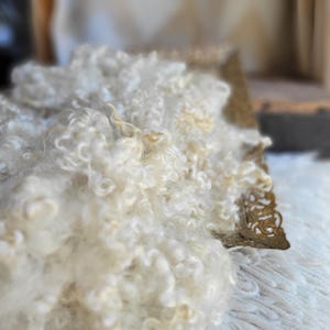 May include: A pile of raw, off-white curly wool fibers with a soft, textured appearance. The wool is resting against a rectangular, ornate, metallic gold-toned tray featuring intricate filigree patterns along its edges and corners.
