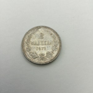 May include: A silver coin with the inscription "2 Markkaa 1872" surrounded by a wreath of oak leaves.