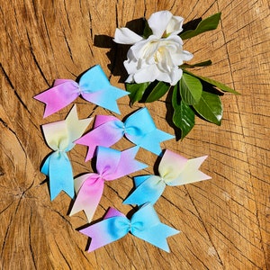 May include: Six ombre hair bows in shades of pink, blue, and yellow. The bows are arranged on a wooden surface with a white flower.