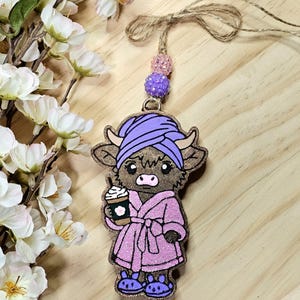 May include: A wooden cow-shaped ornament with a spa theme. The cow wears a purple turban, pink robe, and slippers, holding a coffee cup. The ornament is strung with twine and decorative beads. The background is a light wood surface.