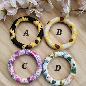 May include: Four different patterned rings, each with a letter label. The rings are black with sunflowers, white with sunflowers, purple and pink floral, and white with green and pink floral.