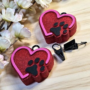 May include: Two heart-shaped pet tag charms with a paw print design. The charms are red with pink trim and black paw prints. Two black metal clips are also in the image. The charms are on a wooden surface with white flowers.