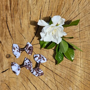 May include: Three white and brown cow print hair bows with a white flower and green leaves on a wooden background. The bows are small and have a classic bow shape.