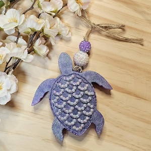 May include: A glittery purple sea turtle ornament with a beaded shell design. The turtle is attached to a string with purple and white beaded accents. Decorative flowers and twine complete the design.