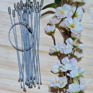 May include: A bunch of silver keychains with a silver loop on a wooden surface. The keychains are made of a thin, braided metal cable. There are white flowers in the background.