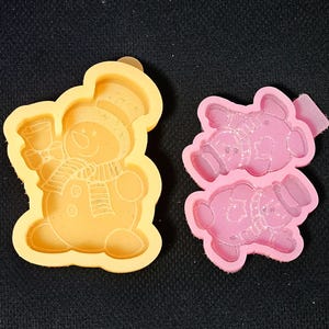 May include: Two silicone molds: one yellow snowman holding a bell, and one pink bear. The snowman mold has a hat, scarf, and buttons. The bear mold has two bears with hats and scarves. Both molds are on a black surface.