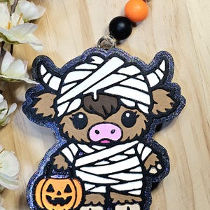 May include: A decorative hanging ornament featuring a brown highland cow dressed as a mummy in white bandages, holding an orange jack-o'-lantern bucket. The figure has a dark purple glittery border and is attached to a jute string with orange and black beads.