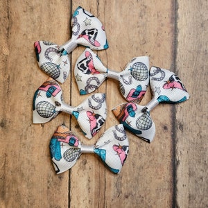 May include: Six white fabric hair bows with a colorful western-themed print. The print includes stars, horseshoes, cowboy hats, and disco balls. The bows are tied with silver ribbon.