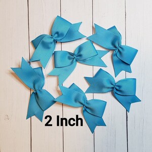 May include: Seven turquoise ribbon bows, each 2 inches in size, are arranged on a light wood-grain surface. The bows have a classic bow shape with a ribbed texture. The text "2 Inch" is displayed in black.