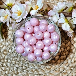 May include: A clear glass bowl filled with light pink, round beads. The beads have a pearlescent sheen and a small hole through the center. White and pink flowers are in the background, with a woven, tan surface beneath the bowl.