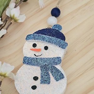 May include: A snowman ornament with a glittery finish. The snowman has a blue hat and scarf, black eyes and buttons, and an orange carrot nose. The ornament is hanging from a string with blue and white beads. The background is a light wood surface.