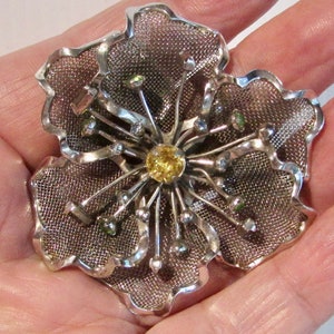 May include: A silver mesh flower brooch with a yellow centre stone and green accents.