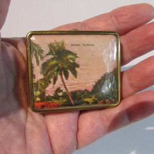 May include: A small, rectangular, brass-framed picture with a vintage postcard image of palm trees and a sunset sky. The postcard reads "MIAMI, FLORIDA."
