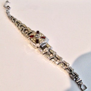 May include: A silver bracelet with a geometric design set with clear and red rhinestones. The bracelet has a clasp closure.