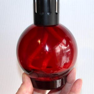 May include: A red glass oil burner with a black metal top and a round shape. The burner has a wick and is designed to hold oil and fragrance.
